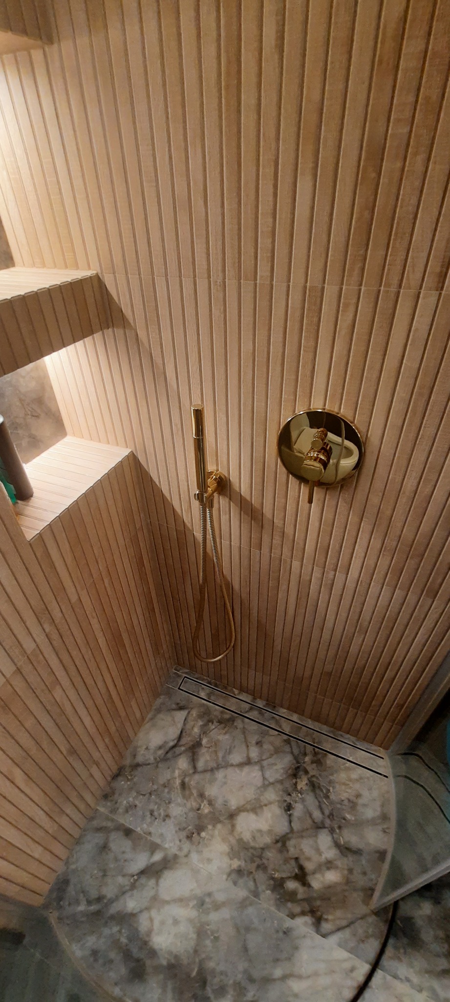 Nowoczesna kabina prysznicowa z okładziną ścienną z drewnopodobnych paneli, złotą armaturą i marmurową posadzką z odpływem liniowym.