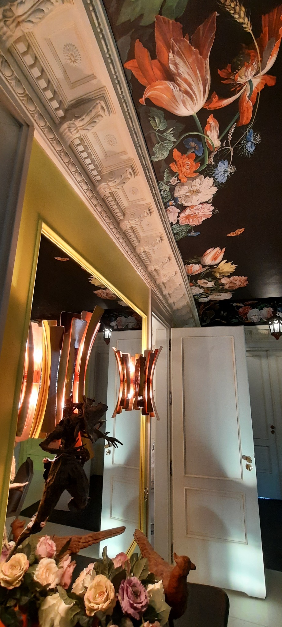 Eleganckie wnętrze z dekoracyjnym malowaniem kwiatowym na suficie, ozdobnym gzymsem, nowoczesnym oświetleniem i aranżacją kwiatową w dolnej części kadru.