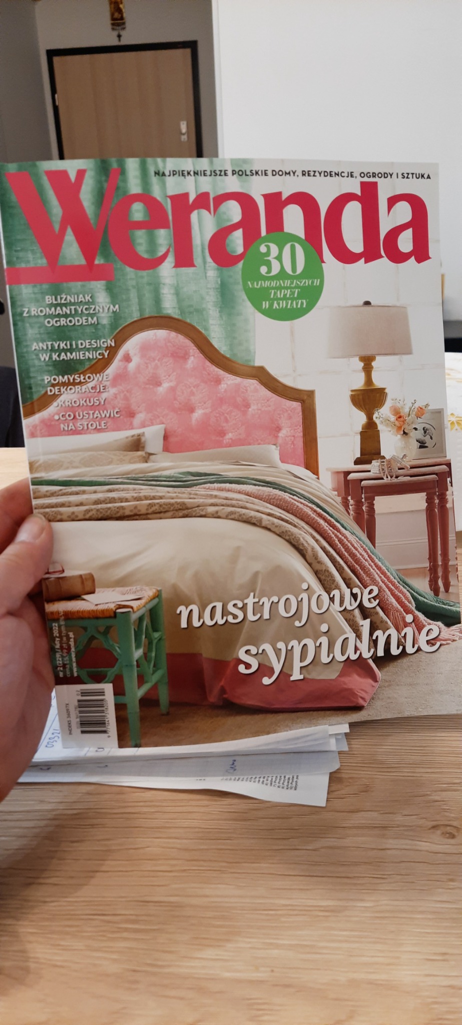 Okładka magazynu 'Weranda' z aranżacją sypialni w pastelowych kolorach, z łóżkiem z tapicerowanym zagłówkiem i drewnianym stolikiem nocnym z lampką. W tle widoczne fragmenty wnętrza.