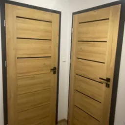 Dwa drewniane drzwi wewnętrzne z czarnymi, poziomymi akcentami i czarnymi ościeżnicami, zamontowane w białej ścianie. Widoczna podłoga z parkietu ułożonego w jodełkę.