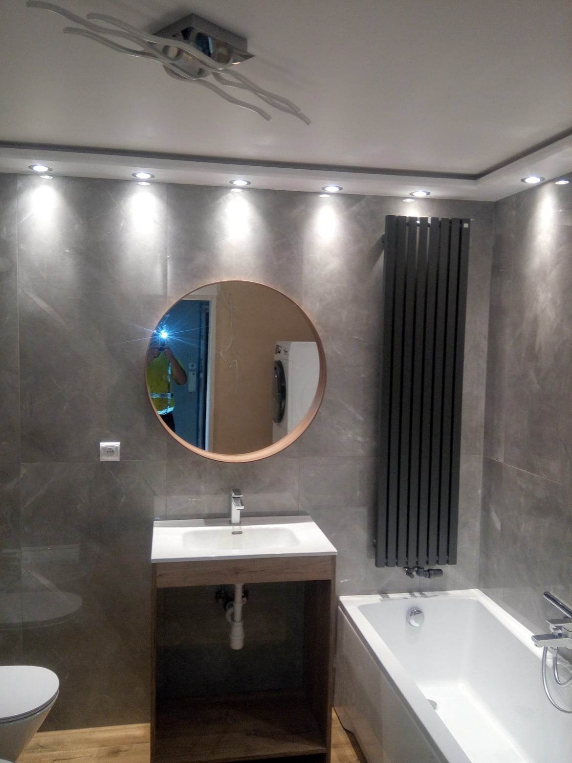 Wykończona łazienka w szarościach z okrągłym lustrem, grzejnikiem panelowym i podwieszanym oświetleniem punktowym. W lustrze odbija się osoba w żółtej kamizelce i pralka.