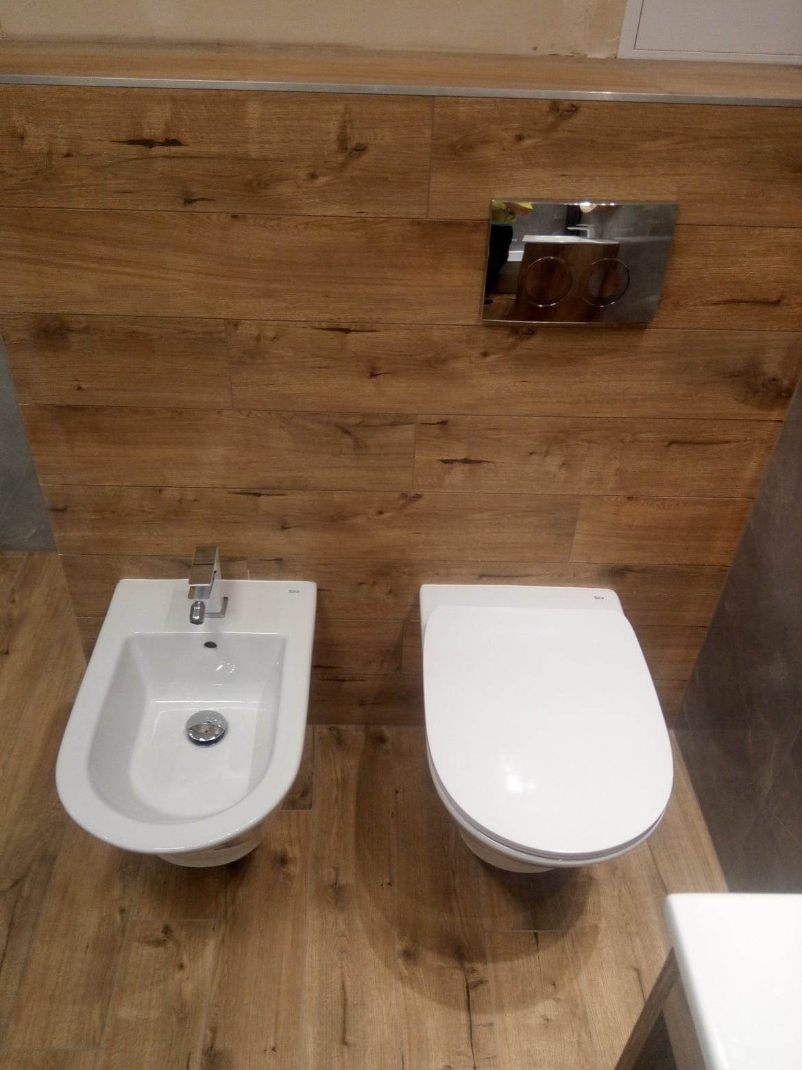 Bidet i wisząca muszla klozetowa w nowoczesnej łazience z drewnopodobnymi płytkami na ścianie i podłodze oraz chromowanym przyciskiem spłukującym.