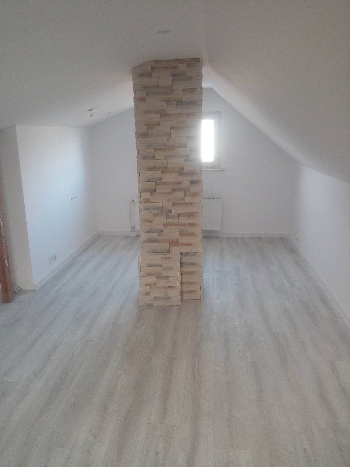 Wykończone poddasze z kolumną obłożoną dekoracyjną, beżową płytką imitującą kamień, na podłodze panele w odcieniach szarości.
