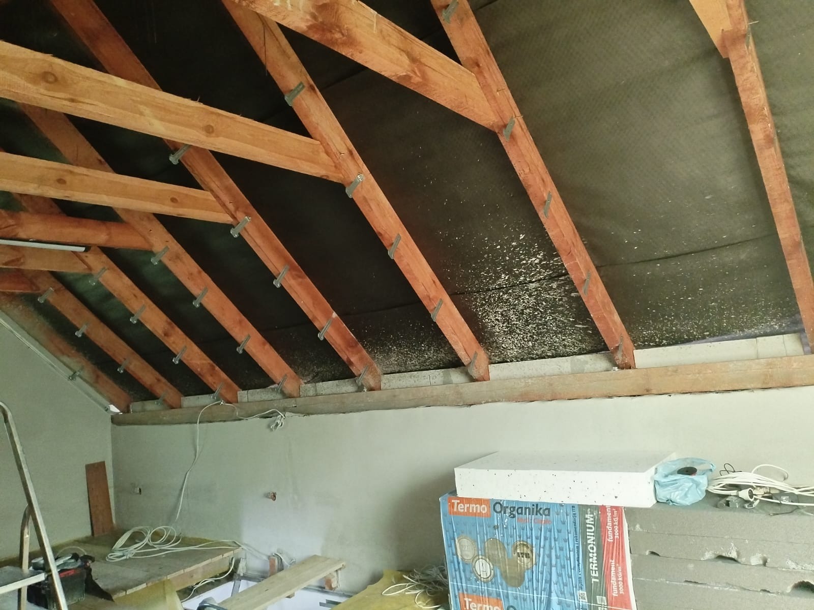 Poddasze w trakcie ocieplania z widoczną konstrukcją drewnianą, czarną folią paroizolacyjną i częściowo zamontowanymi elementami ocieplenia, a także paczkami styropianu Termo Organika na podłodze.