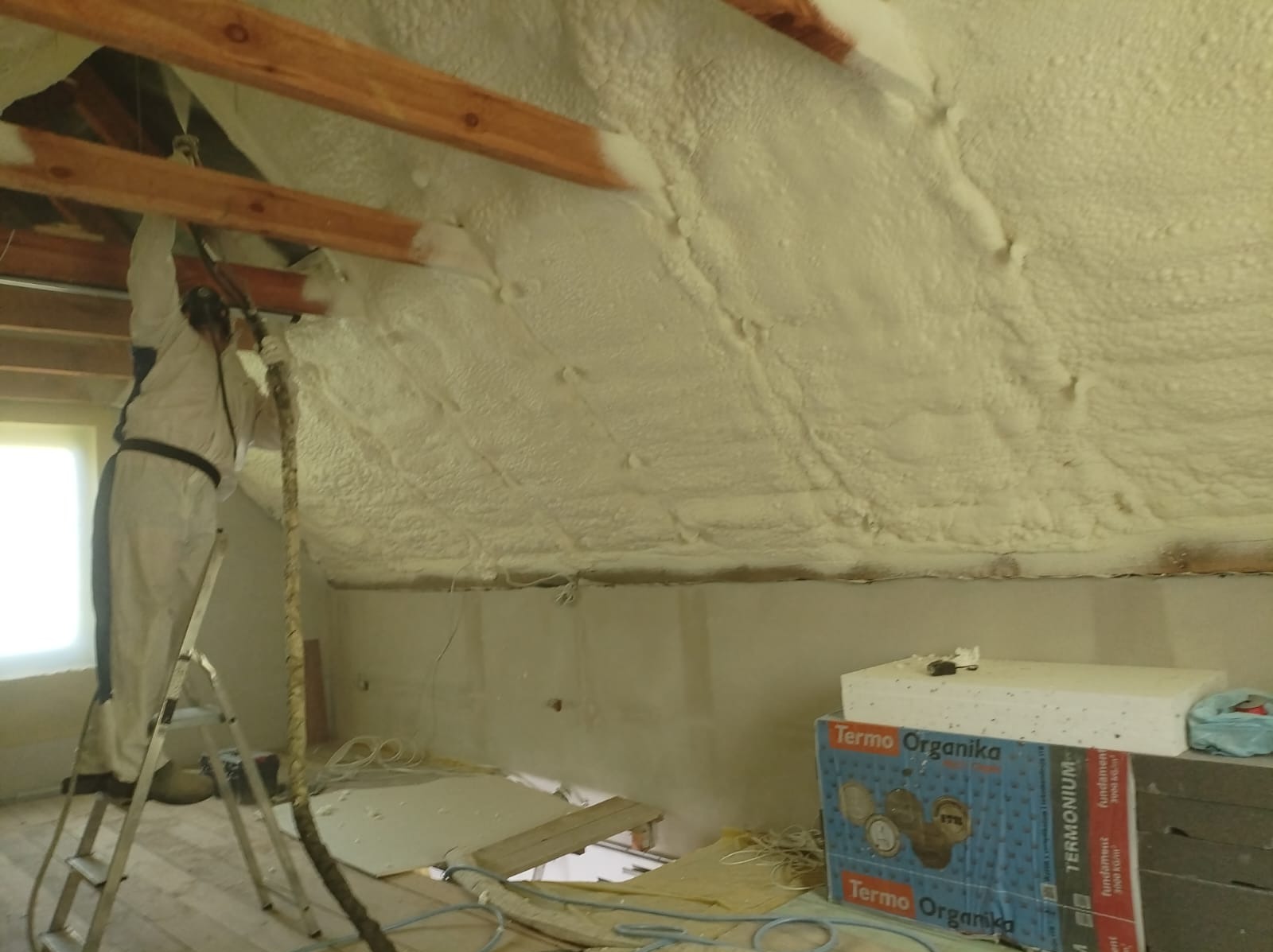 Pracownik w kombinezonie, na drabinie, aplikuje pianę izolacyjną na skośny dach poddasza. Widoczne drewniane belki, częściowo pokryte pianą, oraz kartony styropianu Termo Organika na podłodze.