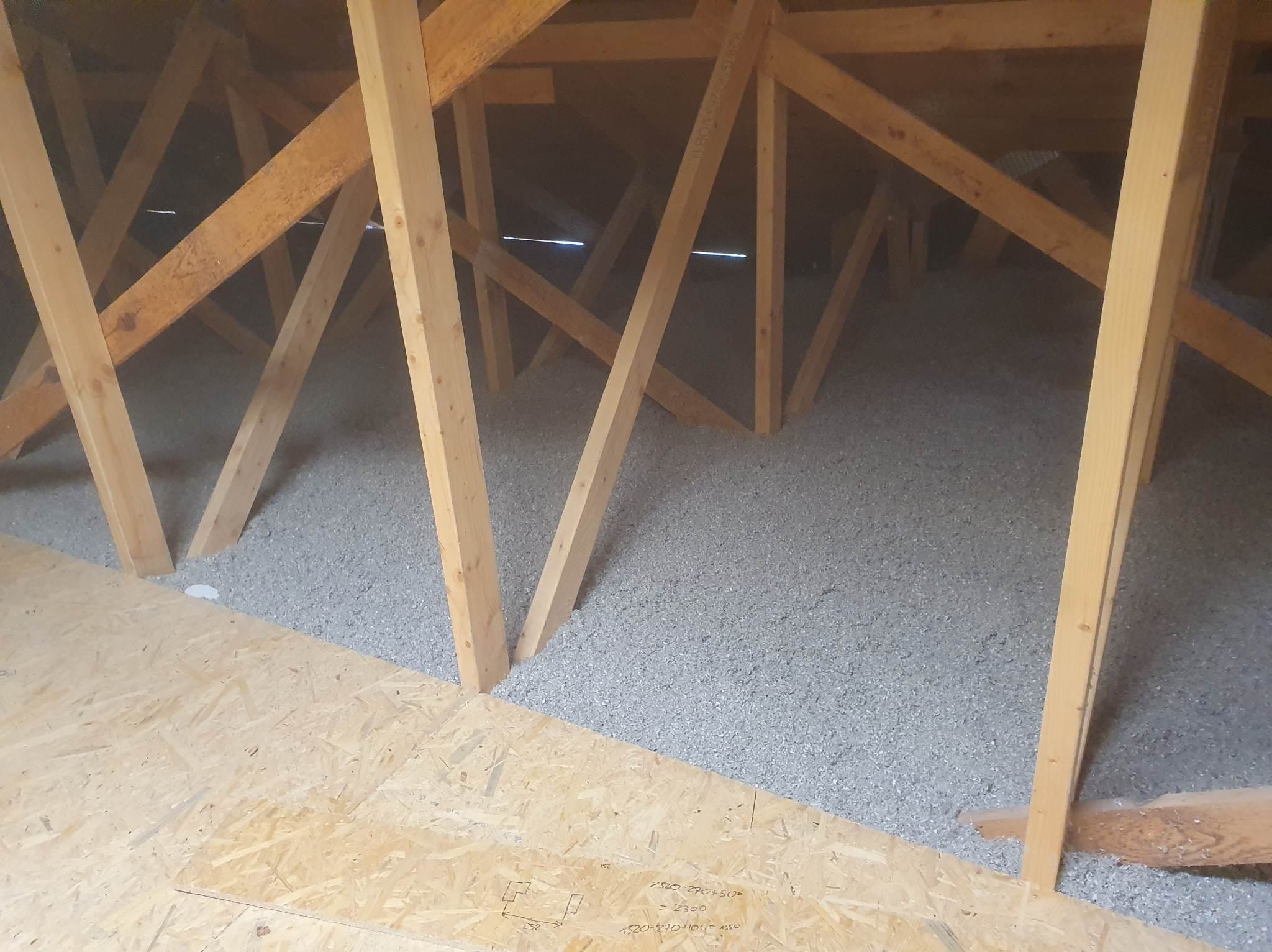 Szara izolacja celulozowa wypełnia przestrzeń poddasza między drewnianymi krokwiami; widoczna płyta OSB z odręcznymi notatkami.