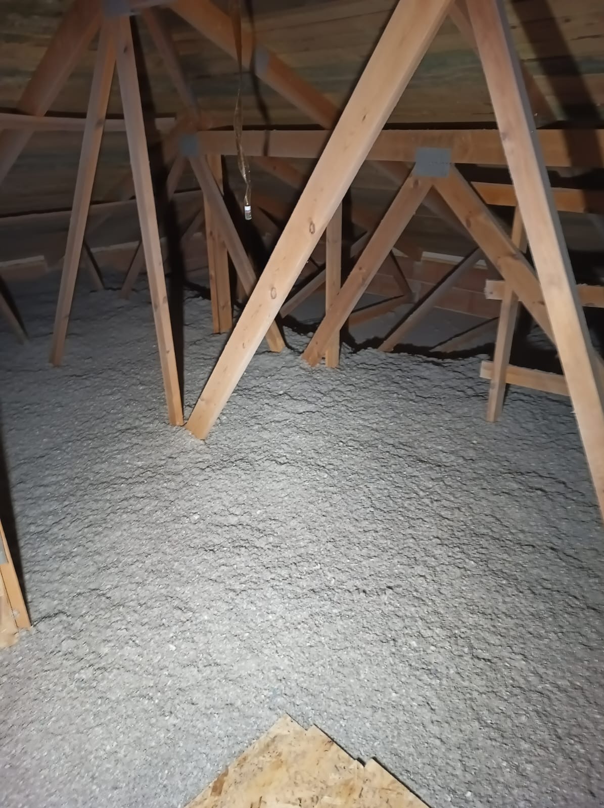 Szara izolacja z wełny mineralnej wypełniająca przestrzeń poddasza, widoczne drewniane elementy konstrukcji dachu, lekko przyciemnione oświetlenie, fragment płyty OSB w rogu kadru.