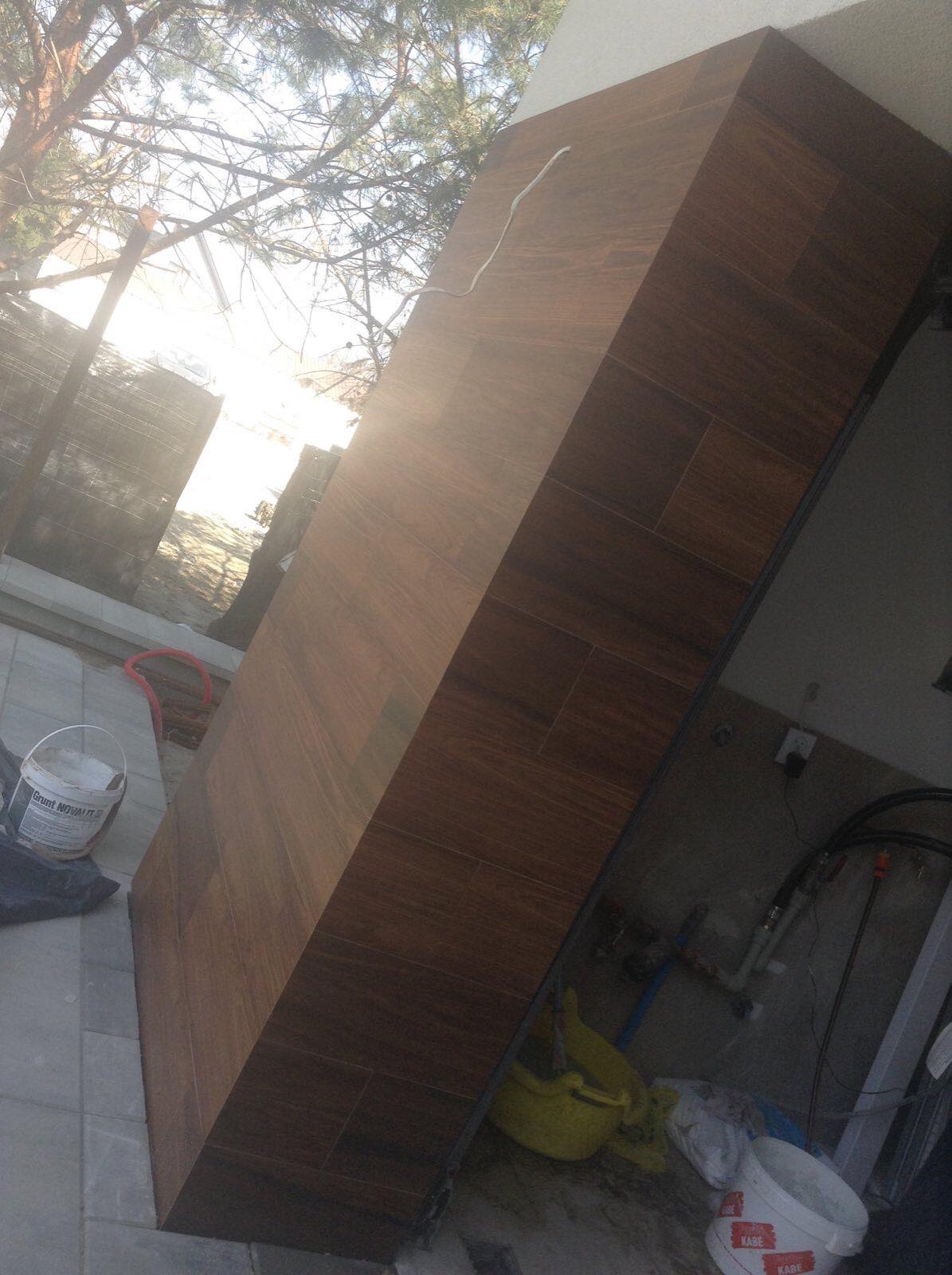 Fragment ściany zewnętrznej budynku wykończony brązowymi płytkami imitującymi drewno; w tle widoczne drzewa i niebo, a także wiadro z farbą i narzędzia.