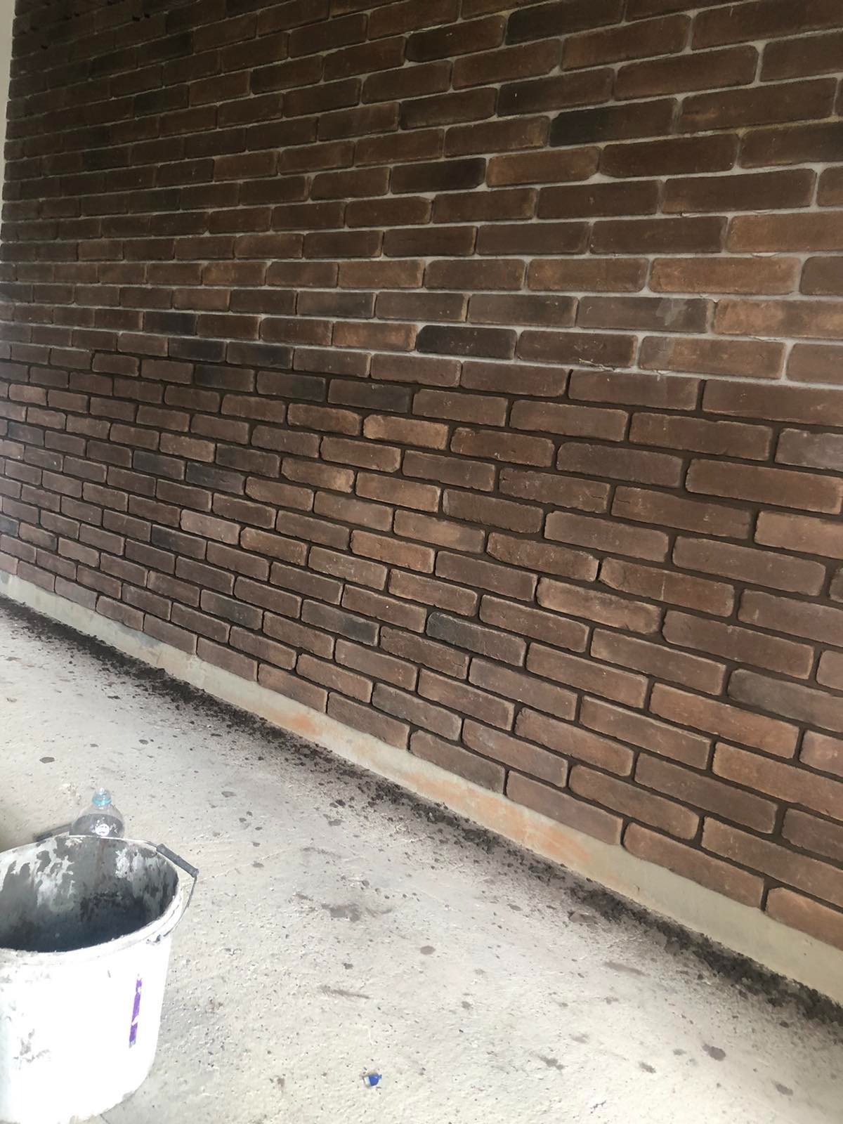 Ściana wyłożona brązową cegłą klinkierową, widoczny fragment podłogi z resztkami zaprawy i wiadro z narzędziami, sugerujące prace wykończeniowe.