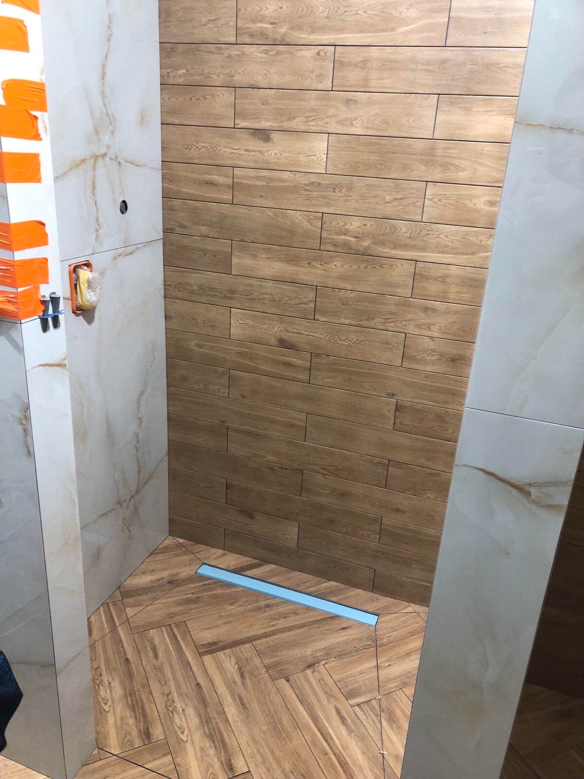 Fragment łazienki w trakcie wykańczania, ściany wyłożone płytkami imitującymi drewno i marmur, podłoga z płytek ułożonych w jodełkę, widoczny odpływ liniowy i zabezpieczone narożniki.