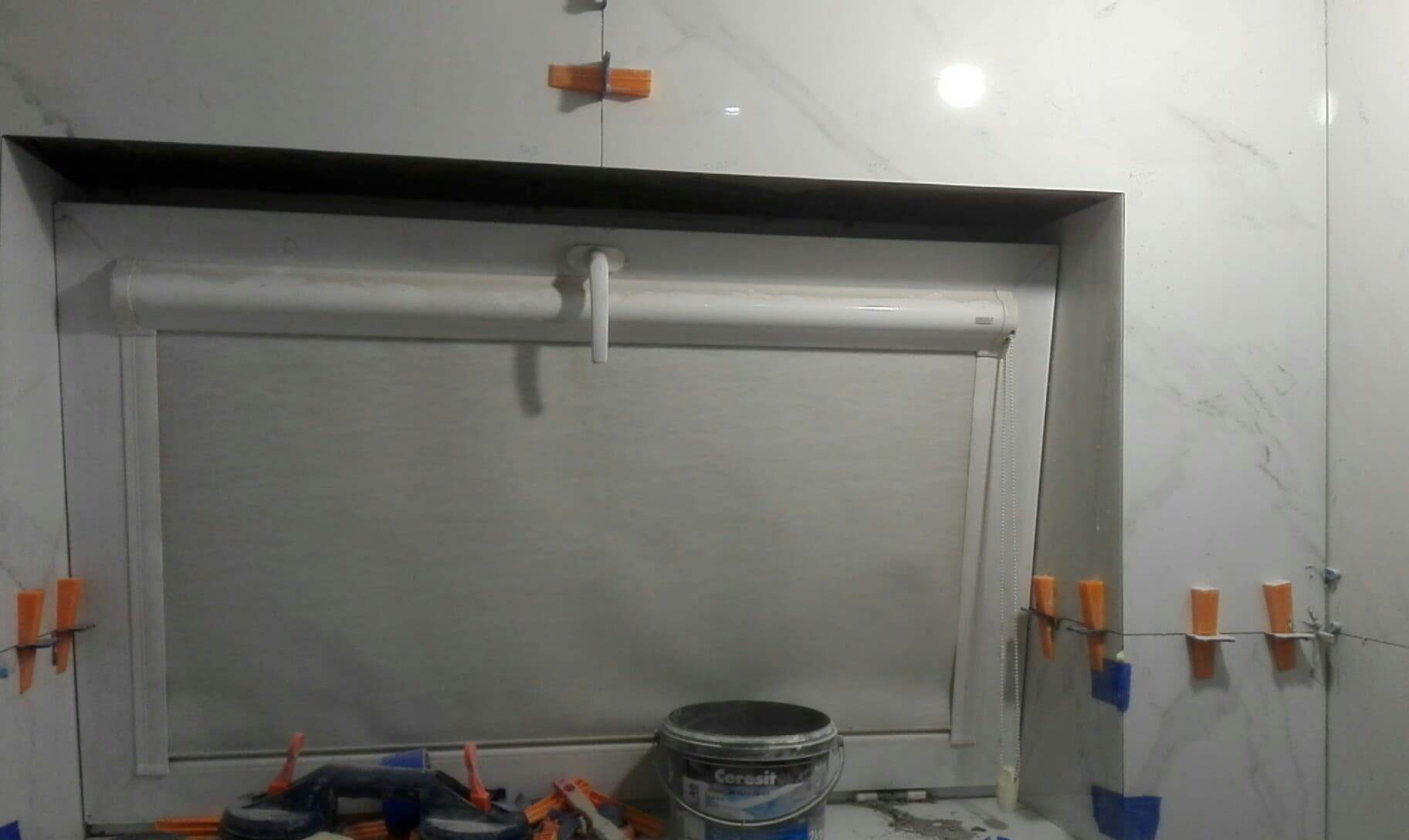 Montaż białych płytek imitujących marmur wokół okna z roletą w łazience, widoczne pomarańczowe krzyżyki dystansowe i wiadro z klejem.