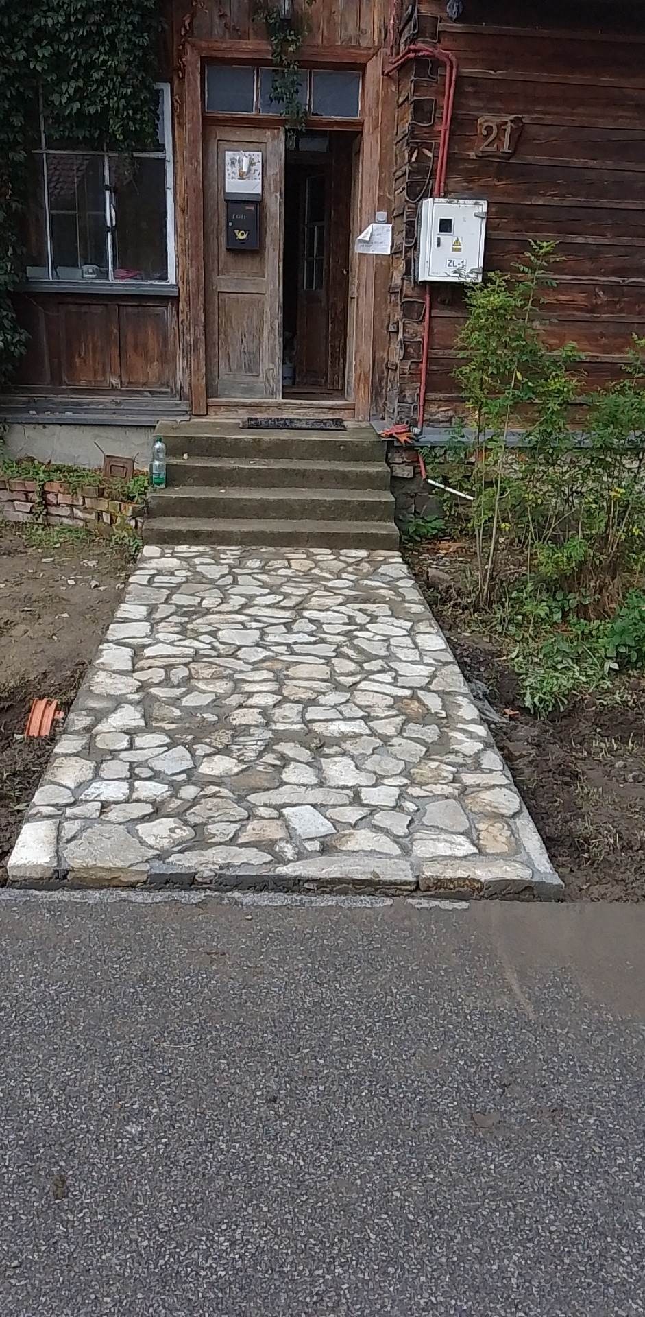 Kamienny chodnik prowadzący do drewnianego domu z numerem 21, widoczne schody i skrzynka pocztowa przy drzwiach.