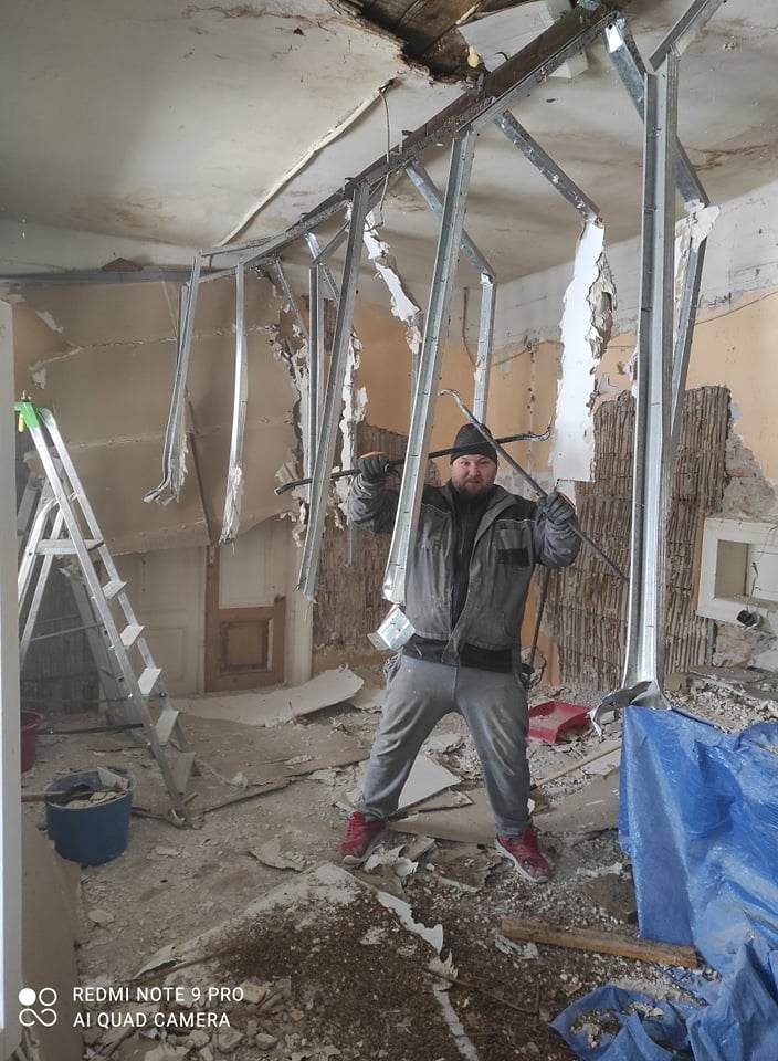 Demontaż konstrukcji sufitu podwieszanego z płyty karton-gips; widoczne metalowe profile, resztki płyty, uszkodzona ściana i mężczyzna w roboczym ubraniu z narzędziem.