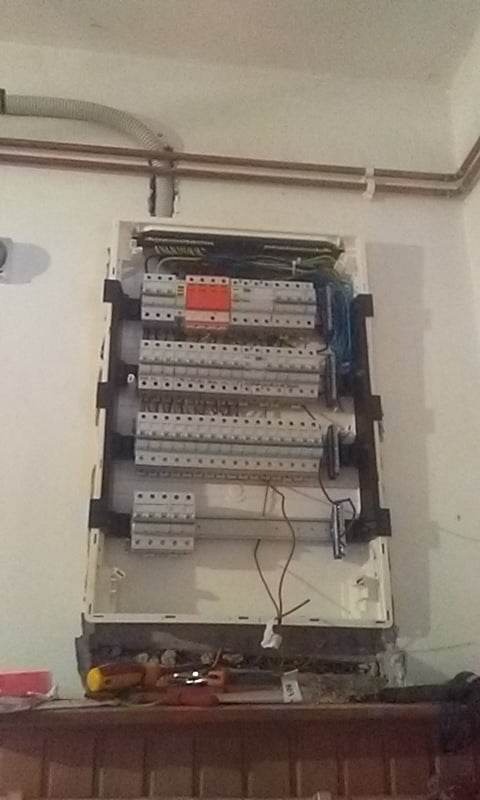 Otwarta skrzynka elektryczna z widocznymi bezpiecznikami, przewodami i narzędziami na półce poniżej, rury na ścianie powyżej.