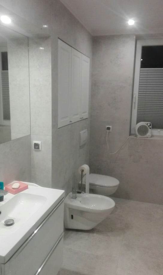 Jasna łazienka po remoncie z umywalką, bidetem i toaletą, wykończona w odcieniach szarości z szafką wiszącą nad toaletą i małym grzejnikiem elektrycznym na parapecie okna.