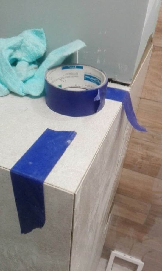 Przygotowanie powierzchni do malowania: rolka niebieskiej taśmy malarskiej i ściereczka leżą na zabezpieczonej taśmą szafce, widoczne płytki podłogowe.