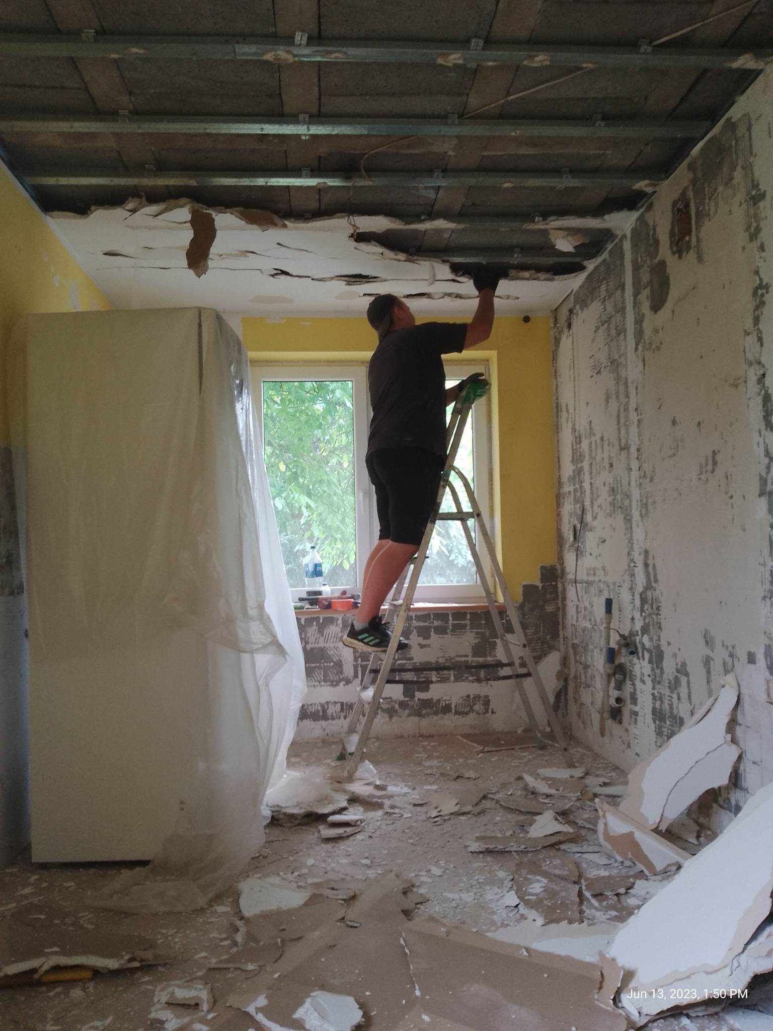 Pracownik na drabinie usuwa resztki starego sufitu podwieszanego, widoczne odsłonięte belki stropowe, zabezpieczona folią szafka, gruz na podłodze, ściany częściowo przygotowane do remontu.