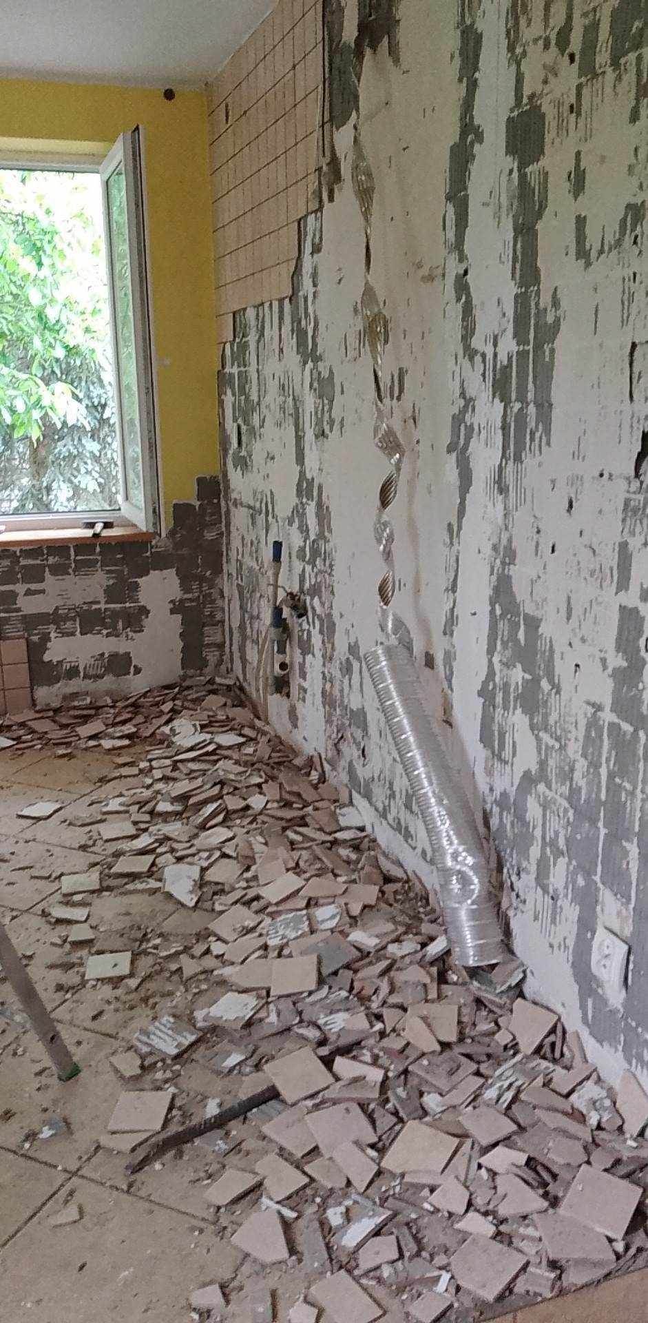 Demontaż starych płytek w łazience, widoczne fragmenty płytek na podłodze, odsłonięta ściana z pozostałościami kleju i instalacją hydrauliczną oraz fragmentem wentylacji.