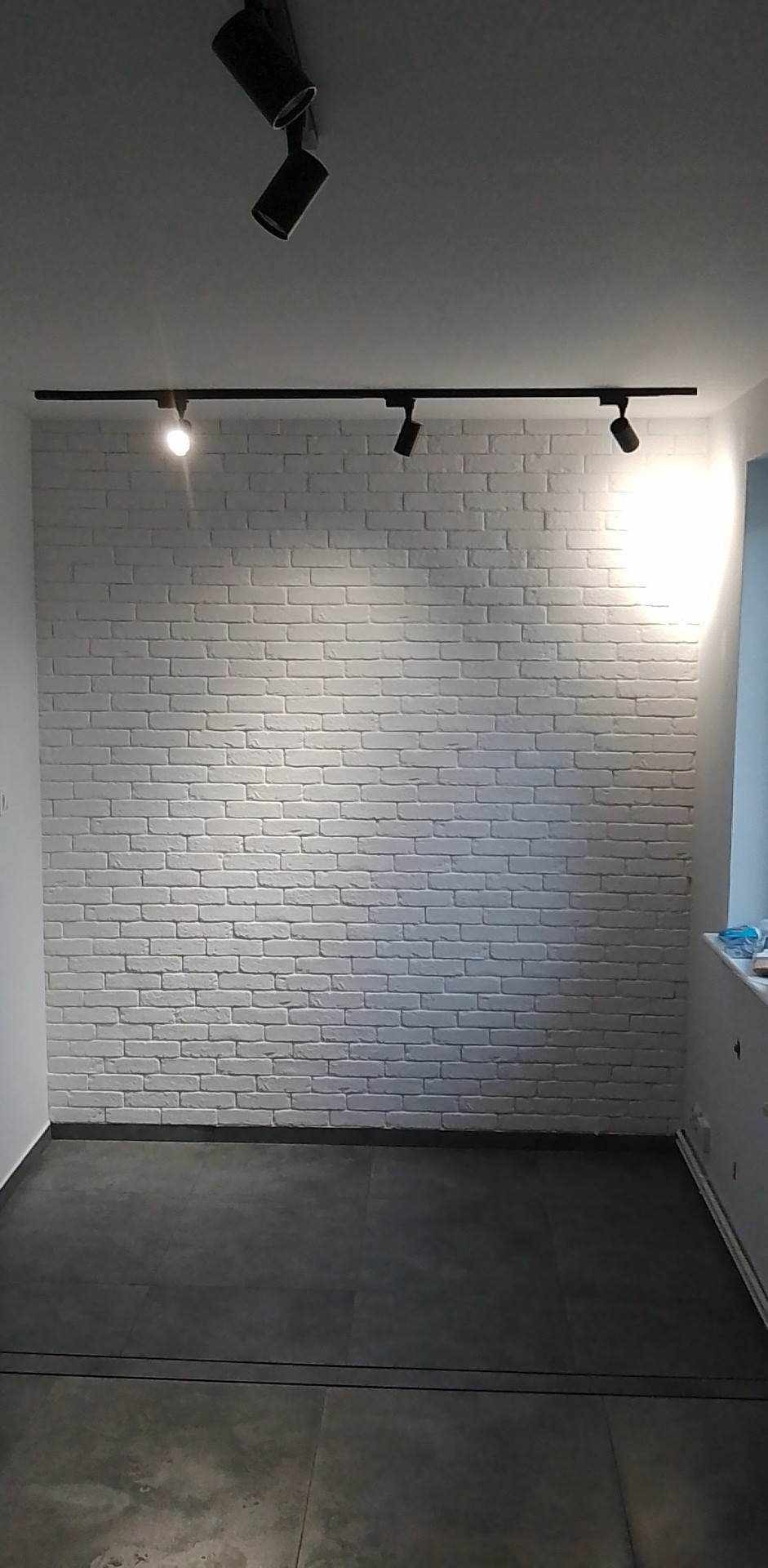 Biała ściana z cegły oświetlona przez czarny system szynoprzewodów z reflektorami, szare płytki podłogowe, widok na fragment okna.