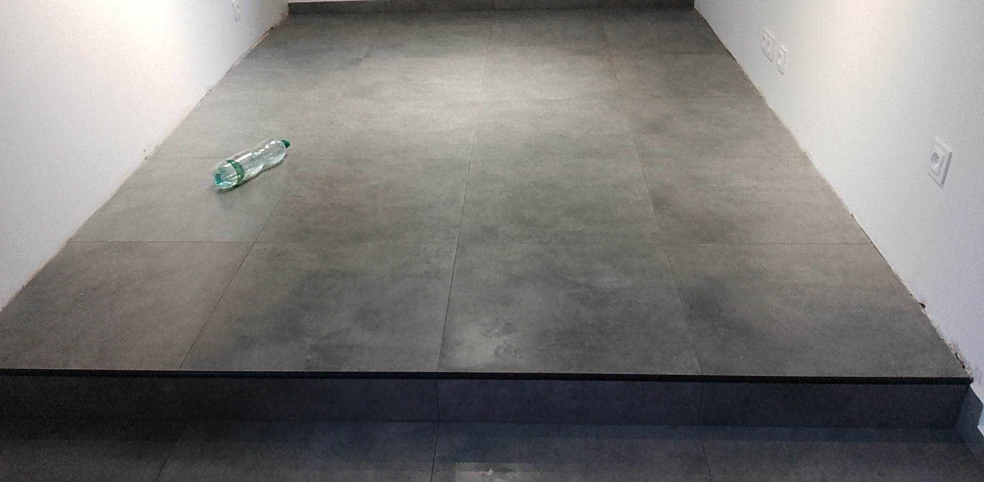 Ułożona szara podłoga z płytek w pomieszczeniu, na podłodze leży pusta plastikowa butelka po wodzie, widoczne gniazdka elektryczne na białej ścianie.