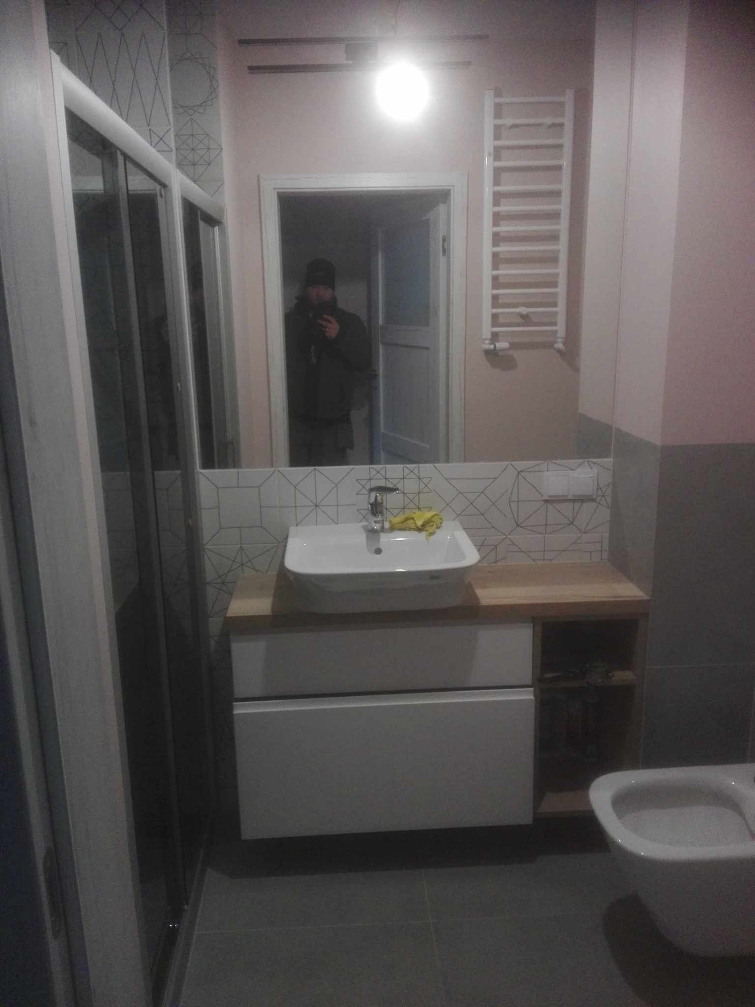 Nowoczesna łazienka po remoncie: biała umywalka nablatowa na drewnianym blacie, geometryczne płytki nad umywalką, grzejnik drabinkowy na ścianie, kabina prysznicowa z przesuwnymi drzwiami po lewej...