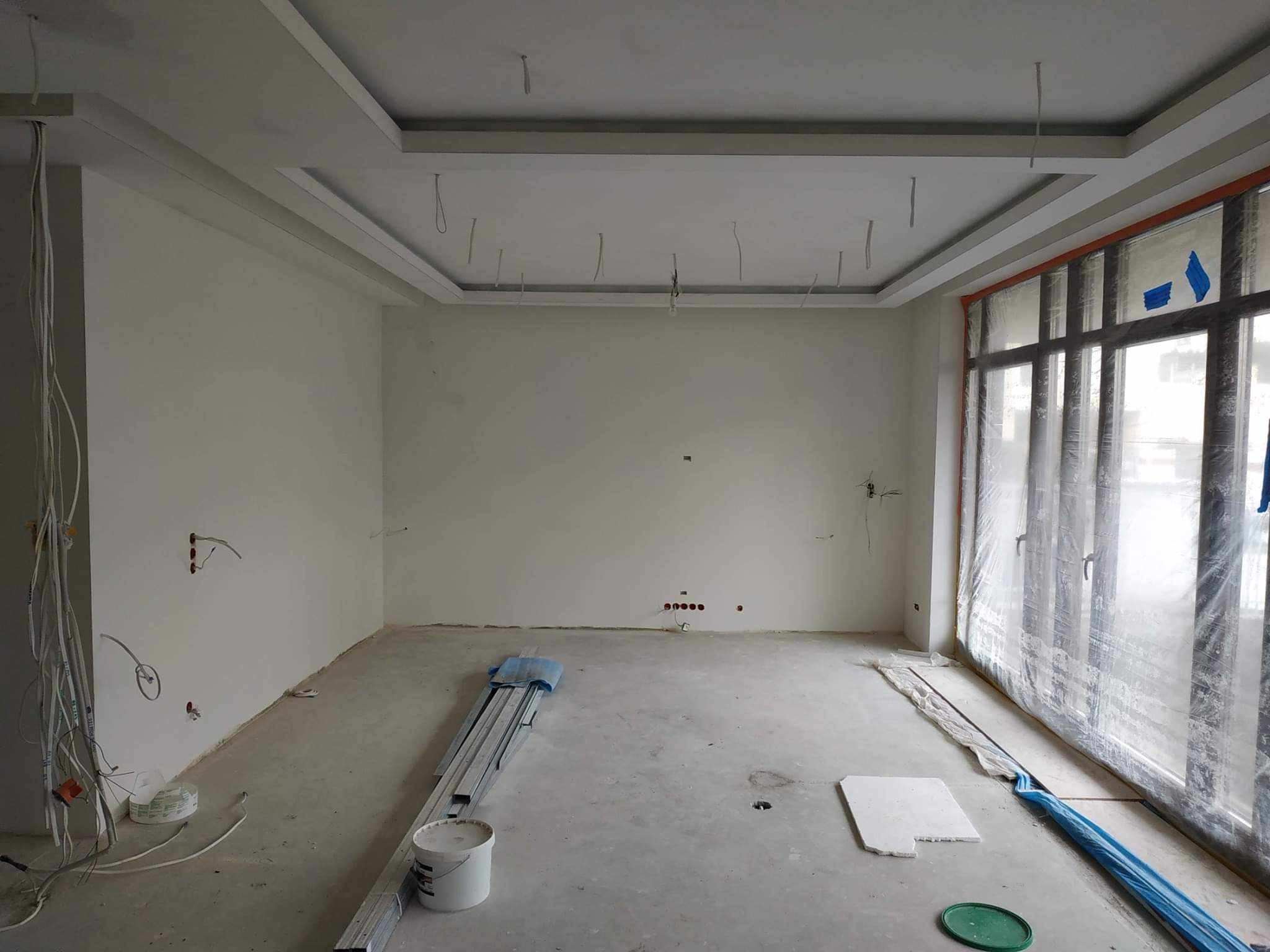 Wnętrze pokoju w trakcie prac wykończeniowych: widoczne przewody elektryczne wystające ze ścian i sufitu, częściowo osłonięte okno, aluminiowe profile i wiadro z farbą na surowej podłodze.