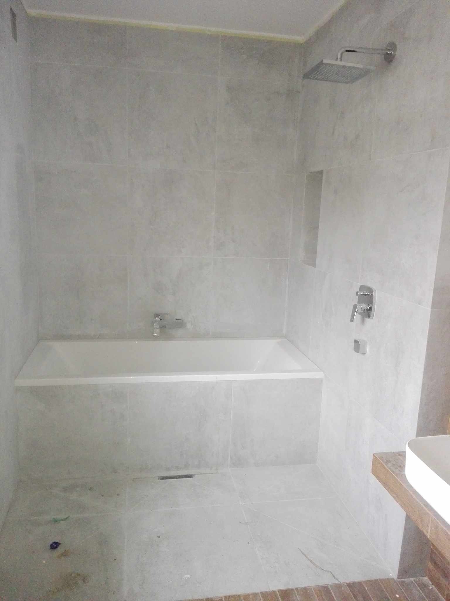 Nowoczesna łazienka w odcieniach szarości z wanną obudowaną płytkami, deszczownicą i minimalistyczną armaturą, widoczne niedociągnięcia wykończeniowe na podłodze.