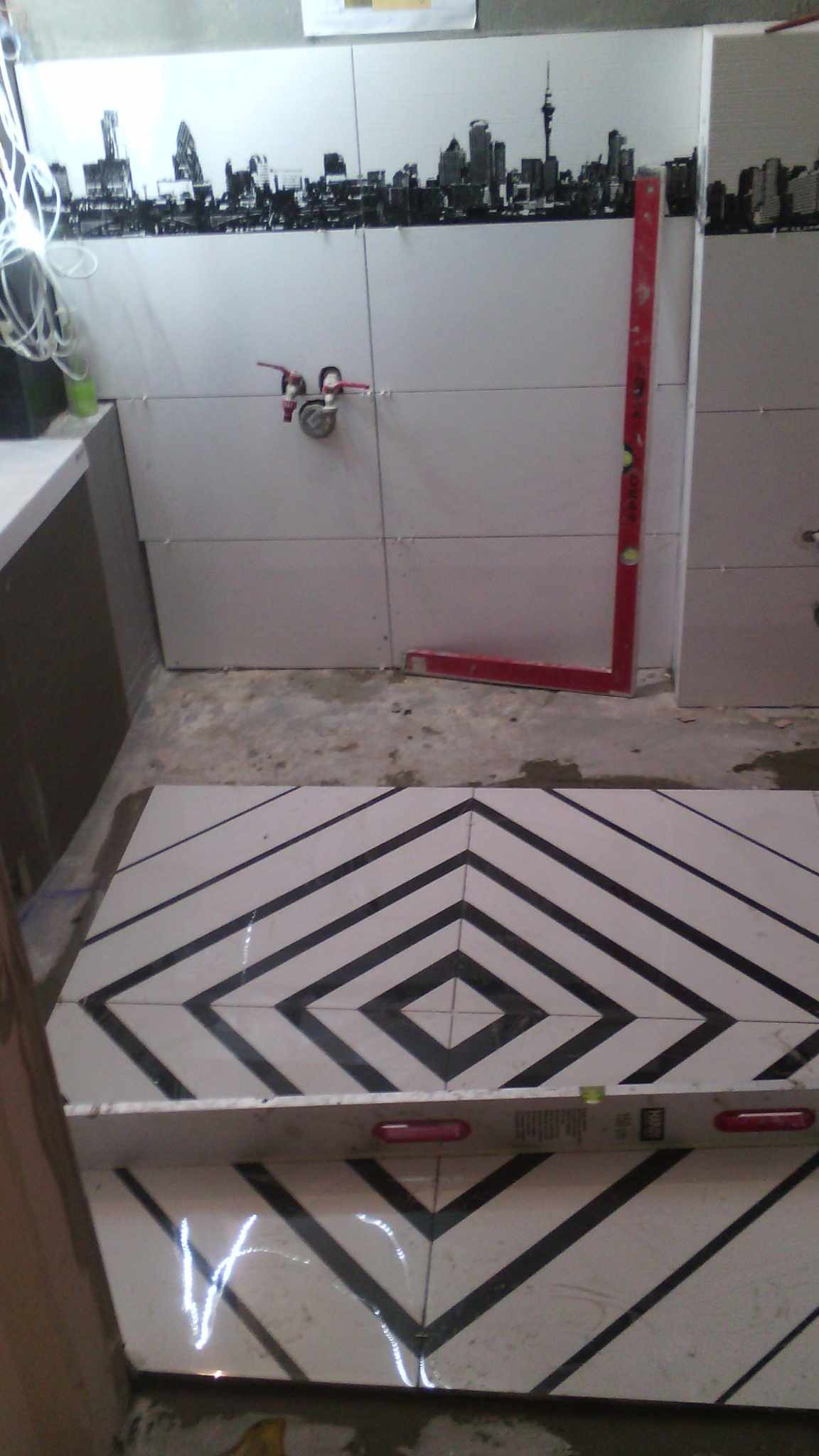 Układanie czarno-białych płytek podłogowych w łazience z motywem rombów, poziomica na płytkach, częściowo ułożone płytki ścienne z motywem panoramy miasta, widoczne narzędzia glazurnicze.