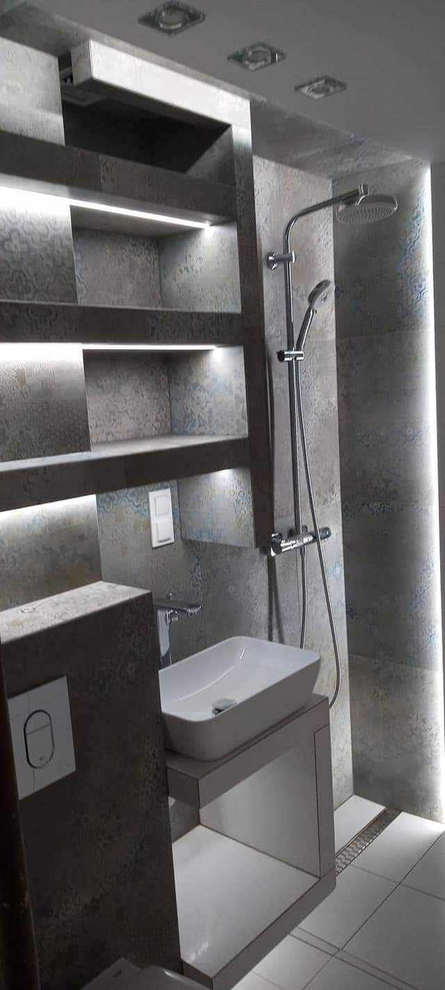 Nowoczesna łazienka z szarymi płytkami imitującymi kamień, podświetlane półki w ścianie, biała umywalka nablatowa i chromowana bateria oraz deszczownica.