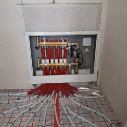 Jarosław Rosiakowski - Rozdzielacz ogrzewania podłogowego z czerwonymi rurami w izolacji, ułożonymi na folii z siatką. Widoczne elementy instalacji w białej szafce w ścianie.