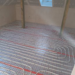 Jarosław Rosiakowski - Instalacja ogrzewania podłogowego w poddaszu z oknem dachowym. Białe rurki ułożone na czerwonej siatce, widoczne drewniane słupy konstrukcyjne.