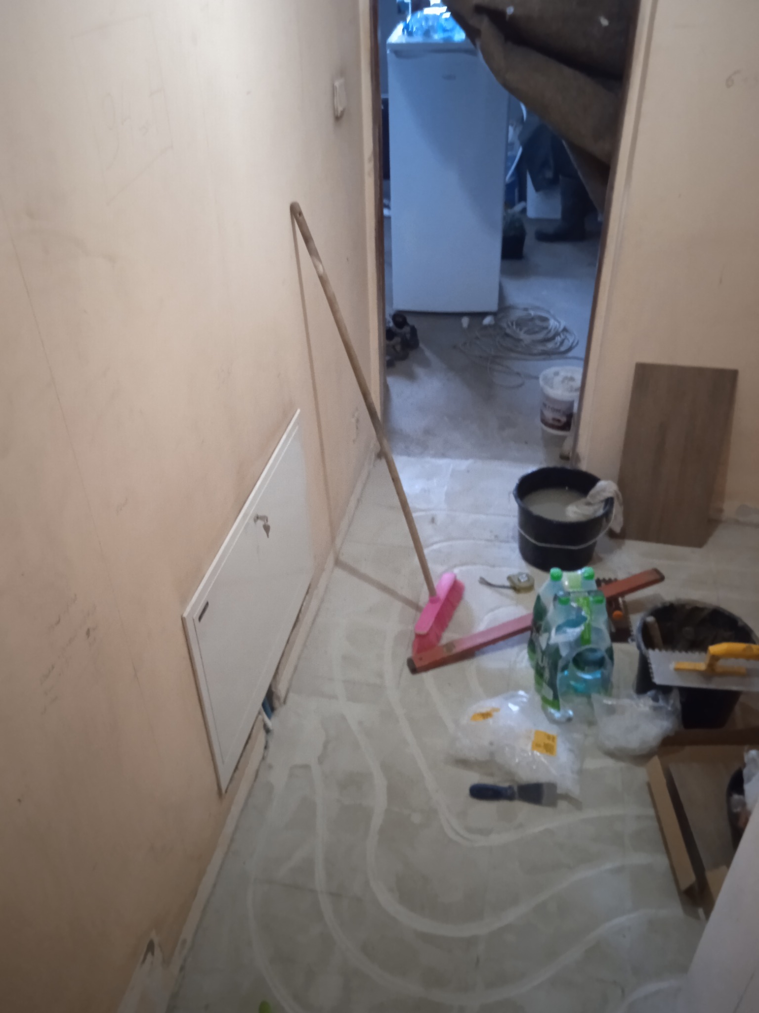 Przygotowanie podłoża pod płytki: gruntowanie posadzki z widocznymi narzędziami, wiadrem z zaprawą i butelkami wody mineralnej w tle remontowanego pomieszczenia.