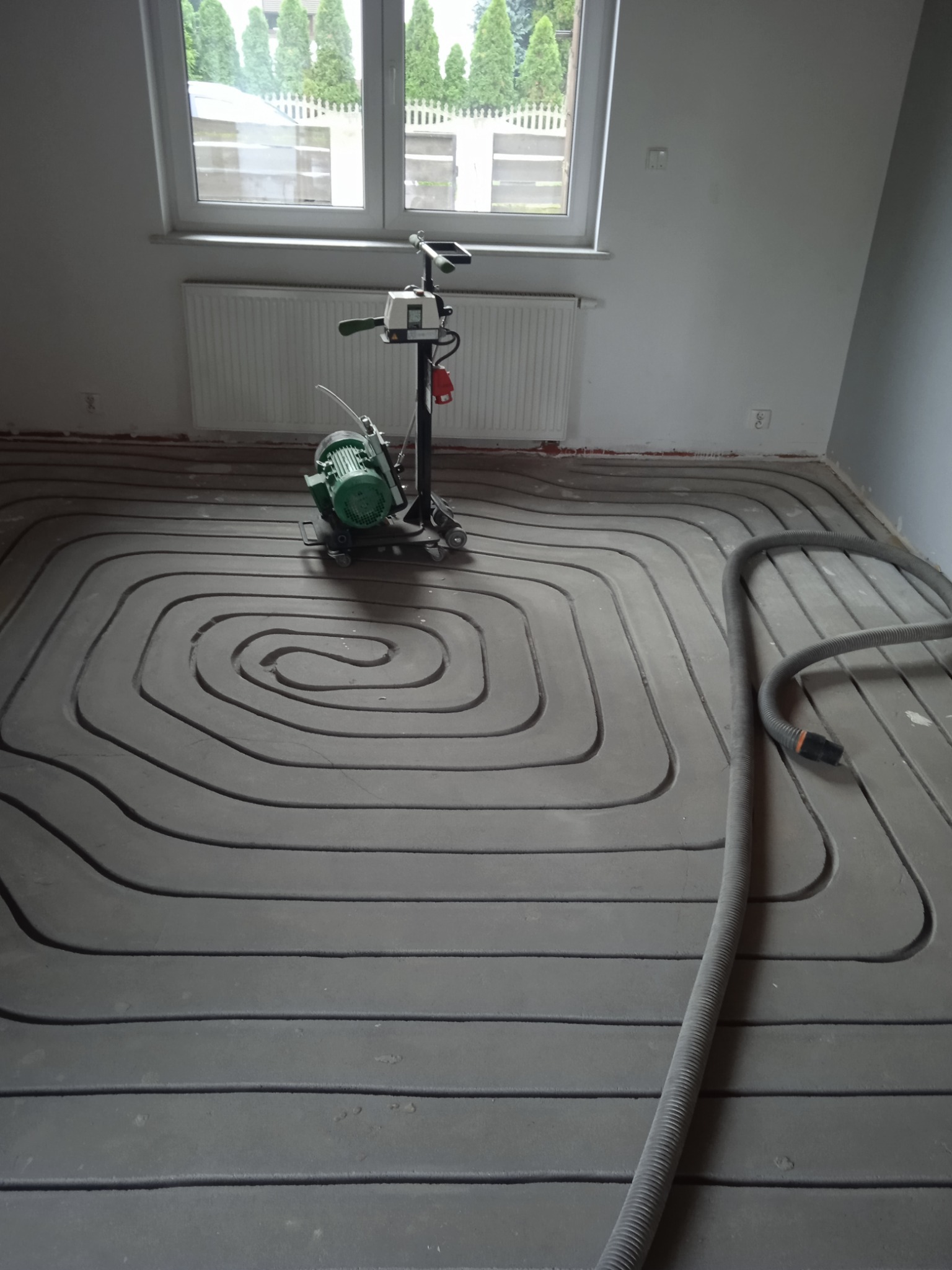 Frezowanie bruzd pod instalację ogrzewania podłogowego w szarym podkładzie, widoczny frezarka i wąż odkurzacza.