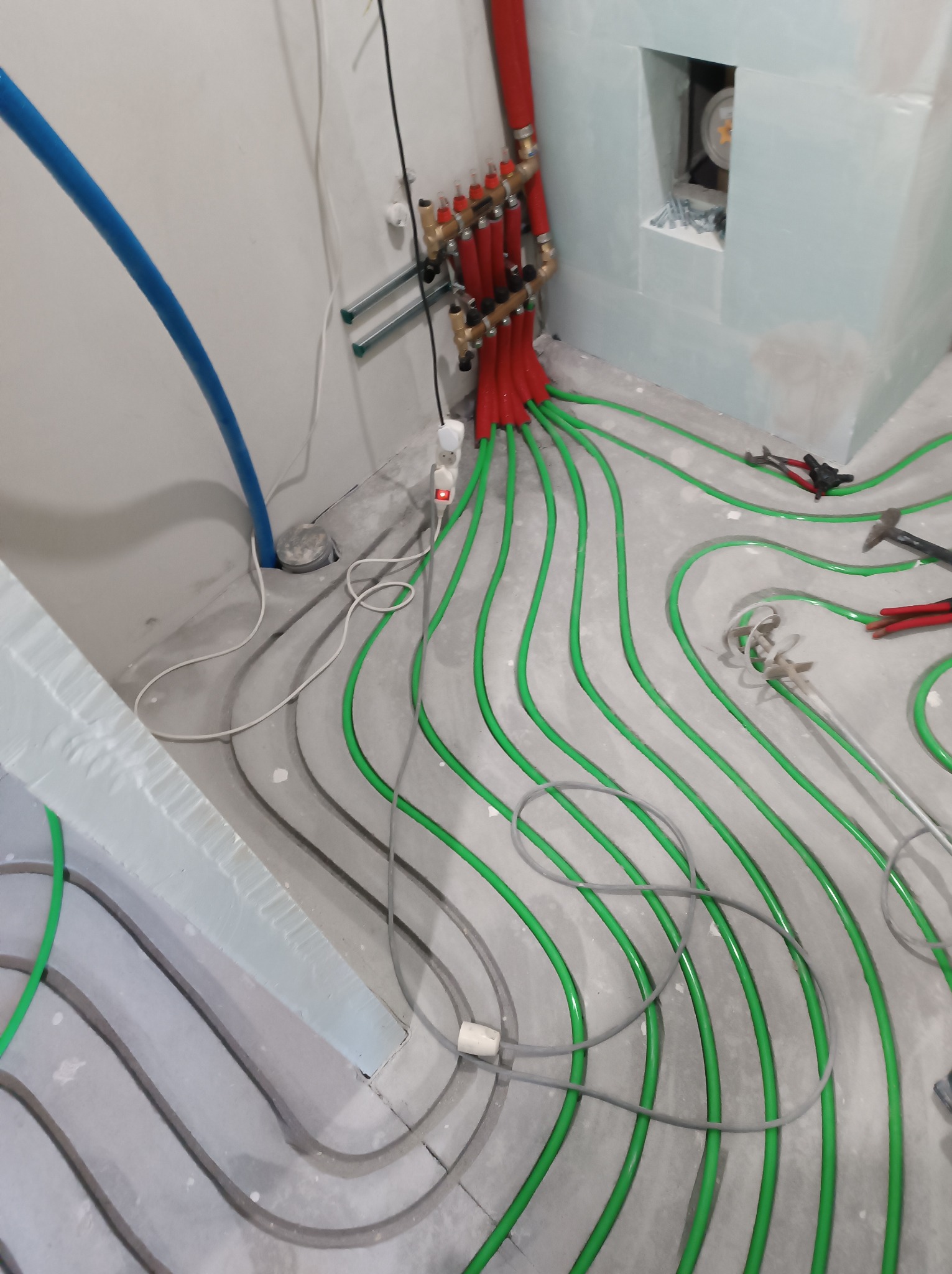 Rozłożone, zielone i szare rury ogrzewania podłogowego na szarym podłożu, z czerwonym rozdzielaczem i narzędziami w tle. Widoczny fragment ściany z płyt karton-gips.