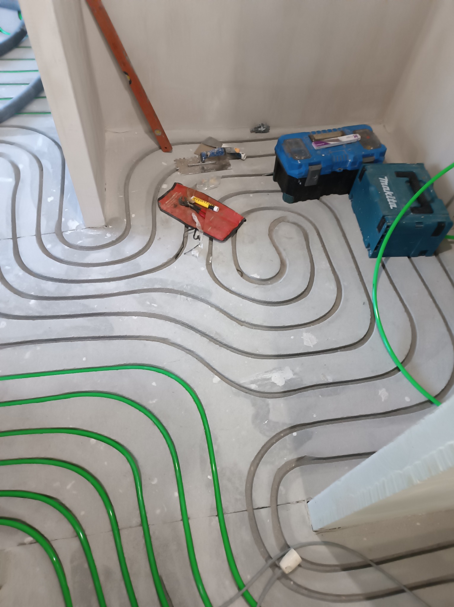 Ogrzewanie podłogowe: zielone rurki ułożone w spiralny wzór na szarym podłożu, narzędzia pracy w tle. Widok z góry na etap instalacji.