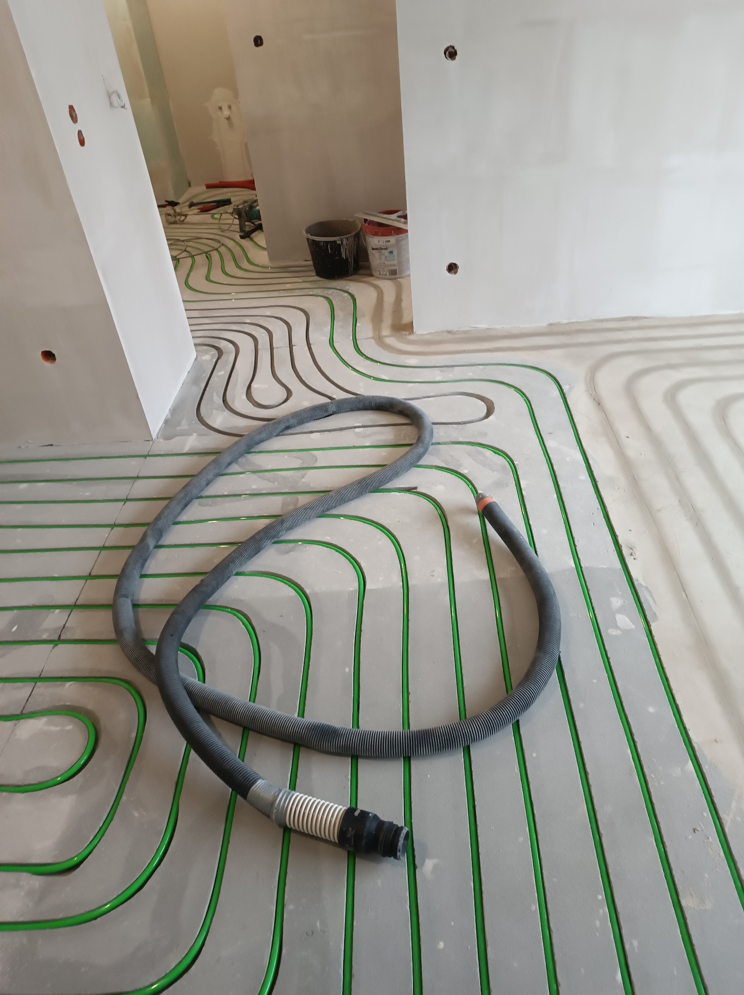 Zielone rurki ogrzewania podłogowego ułożone na szarym podłożu, widoczne przygotowania do wylewki. W tle ściany w surowym stanie i wąż do wylewki.