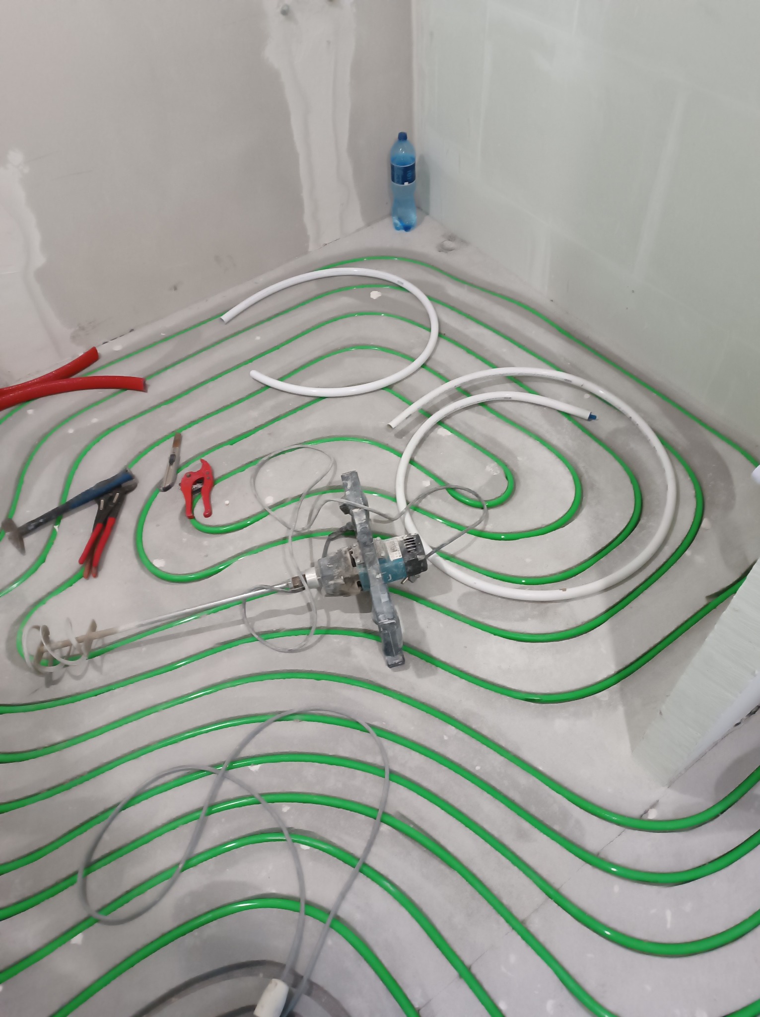 Ułożone, zielone rury ogrzewania podłogowego na szarym podłożu w pomieszczeniu z płytami karton-gips. Widoczne narzędzia i mikser do wylewki. Butelka wody w rogu.