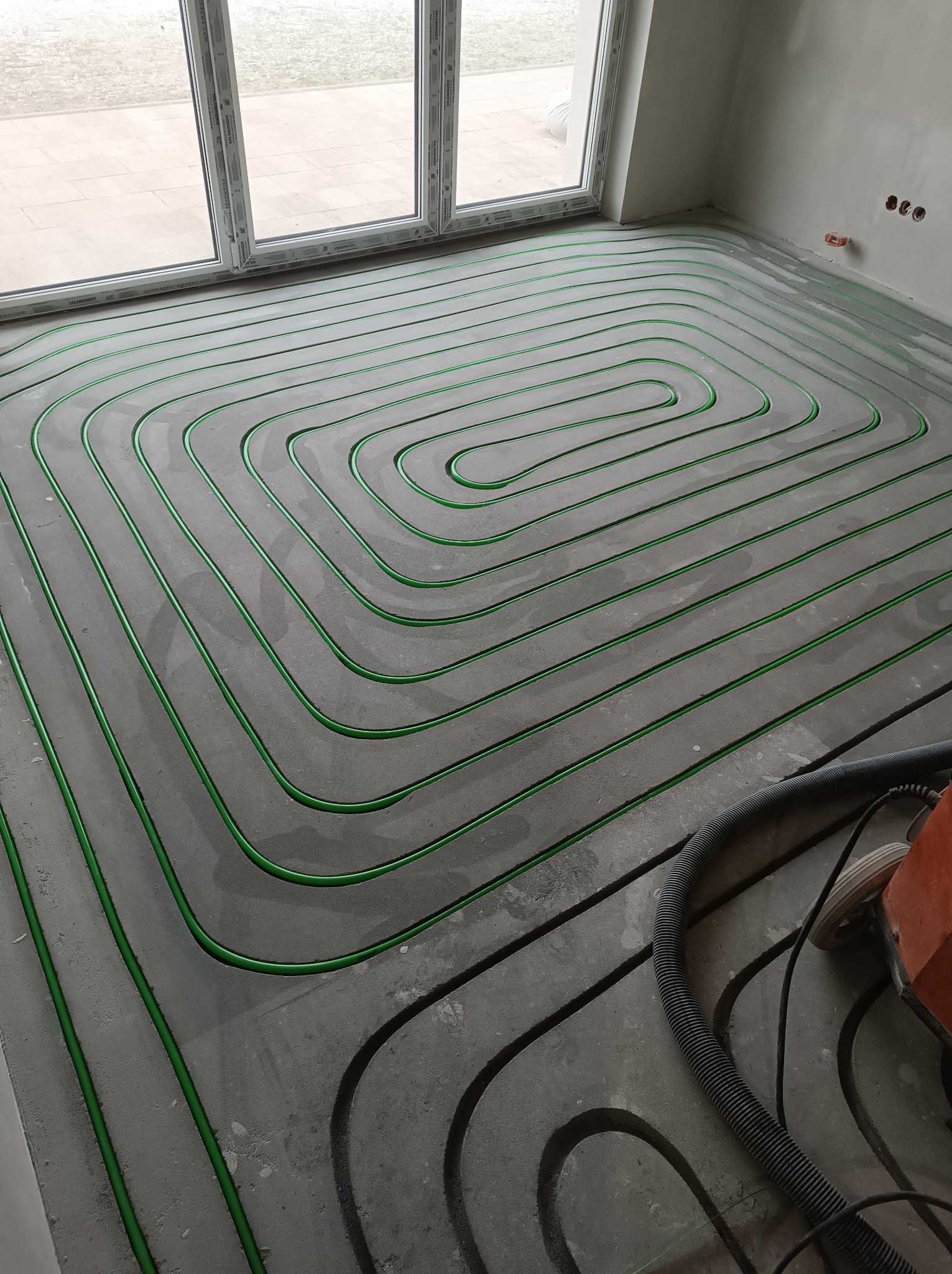 Zielone rurki ogrzewania podłogowego ułożone spiralnie w szarej wylewce, widok z góry, z fragmentem odkurzacza przemysłowego w rogu pomieszczenia.