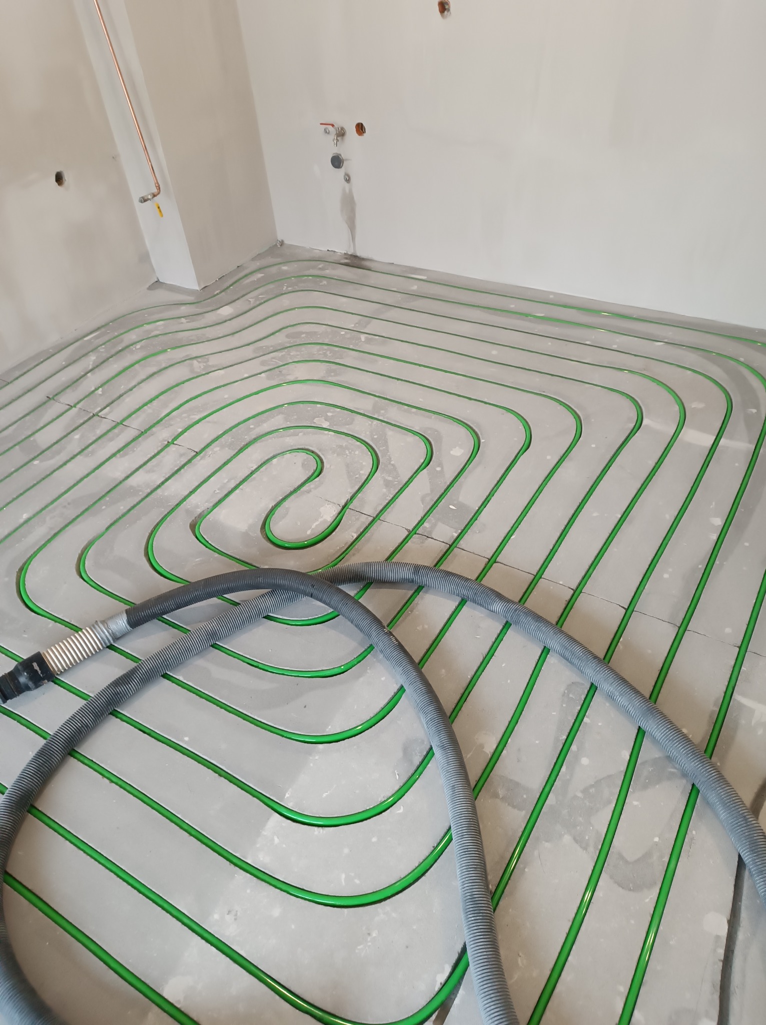 Zielone rurki ogrzewania podłogowego ułożone spiralnie na szarej wylewce, widoczne szare węże do czyszczenia instalacji. Ściany w surowym stanie.