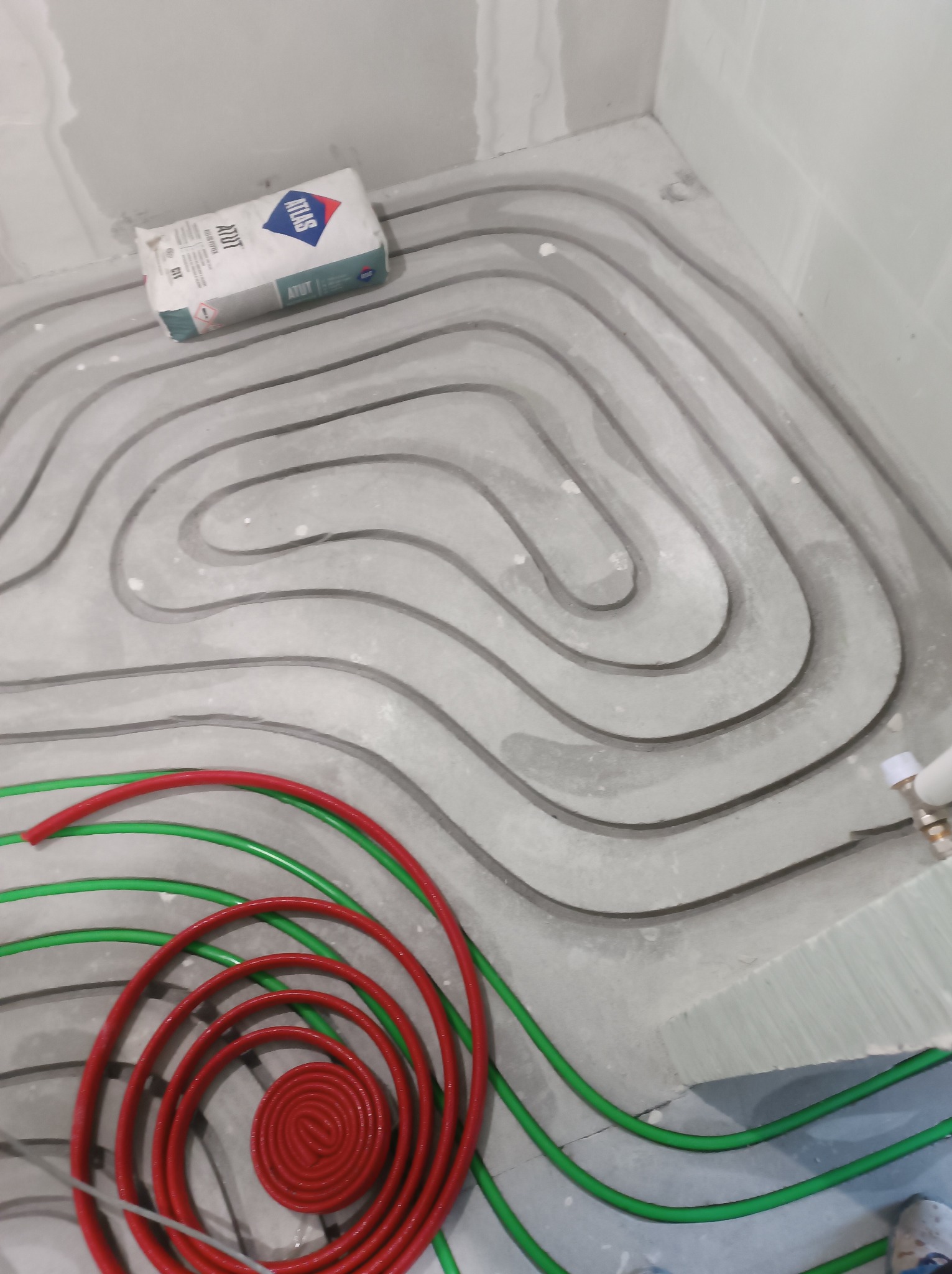 Ułożone rury ogrzewania podłogowego w szarej wylewce, widoczny zwój czerwonej rury i worek zaprawy Atlas. Ujęcie z góry, w trakcie prac instalacyjnych.
