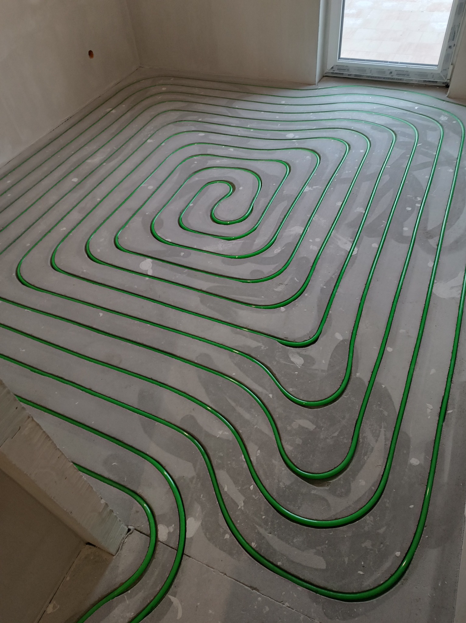 Zielone rury ogrzewania podłogowego ułożone spiralnie na szarym betonie w surowym wnętrzu z oknem. Widok z góry, ukazujący precyzję instalacji.