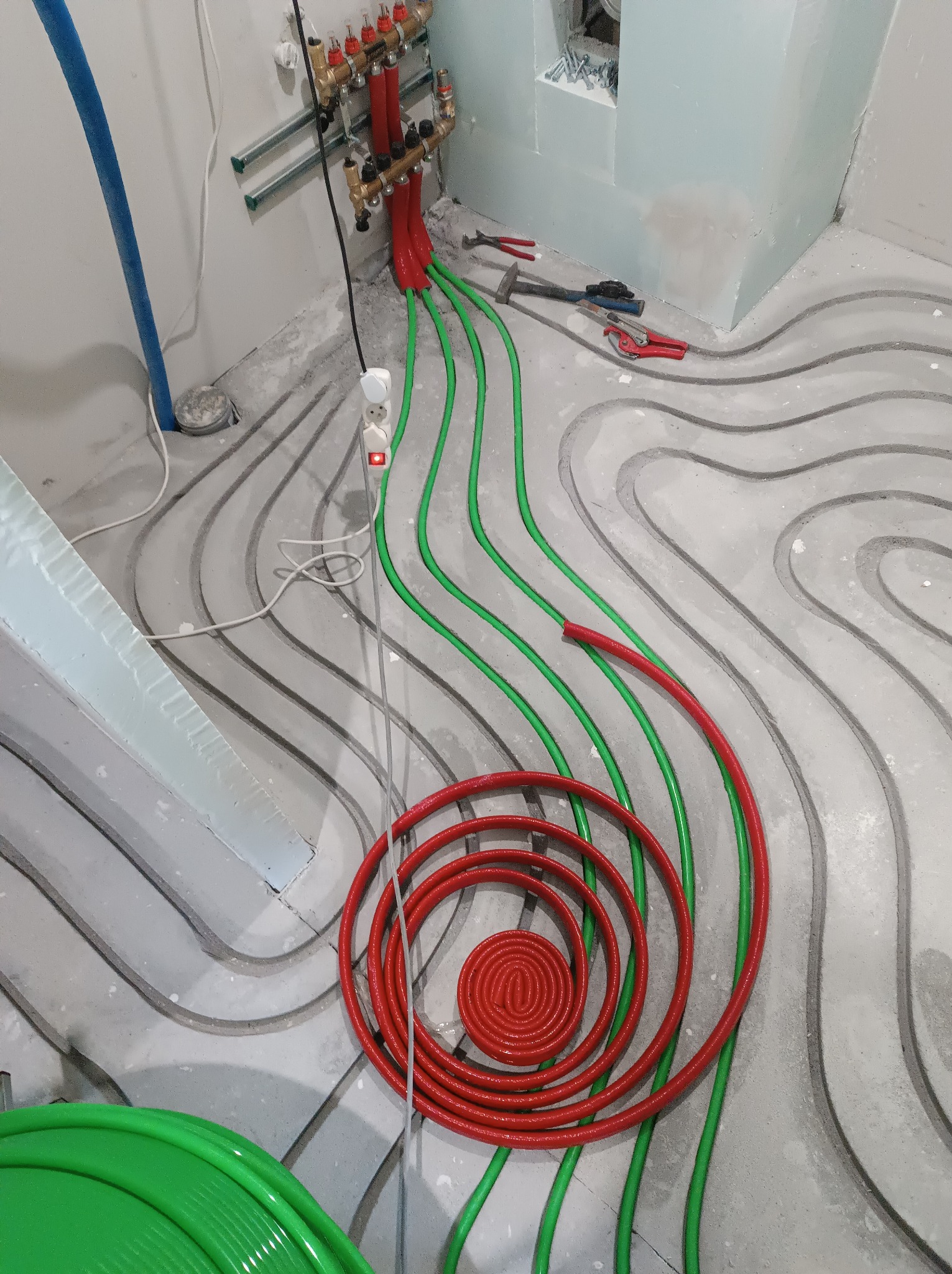 Rozłożona instalacja ogrzewania podłogowego z czerwonymi i zielonymi rurami, widoczne narzędzia i rozdzielacz. Etap prac przed wylaniem posadzki.
