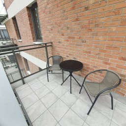 Salvare Serwis - Zestaw mebli balkonowych: okrągły stolik z czarnym blatem i dwa plecione krzesła z metalowymi nogami na jasnej, kwadratowej podłodze z płytek, na tle ściany z czerwonej cegły.