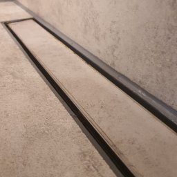 Salvare Serwis - Długi, liniowy odpływ podłogowy w nowoczesnej łazience wykończonej w odcieniach szarości, z widoczną strukturą betonu na ścianach i podłodze.