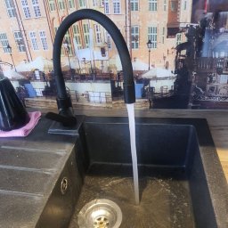 Salvare Serwis - Czarny, elastyczny kran z włączoną wodą nad czarnym, kamiennym zlewem. W tle panel ścienny z motywem starego miasta.