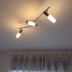 Chromowana lampa sufitowa z trzema regulowanymi reflektorami o cylindrycznym kształcie, oświetlająca pomieszczenie z delikatną firanką w tle.