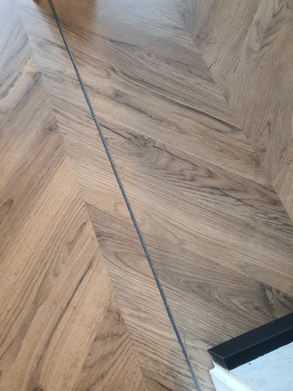 Ułożona w jodełkę podłoga z paneli drewnopodobnych, z widoczną listwą przypodłogową w dolnym rogu.