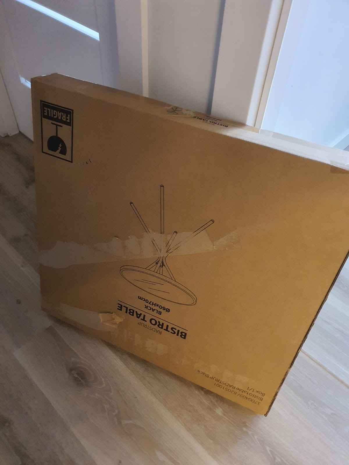 Kartonowe opakowanie płaskiego paczki z nadrukowaną grafiką czarnego stolika bistro Radstrup, widoczny znak ostrzegawczy 'Fragile'.