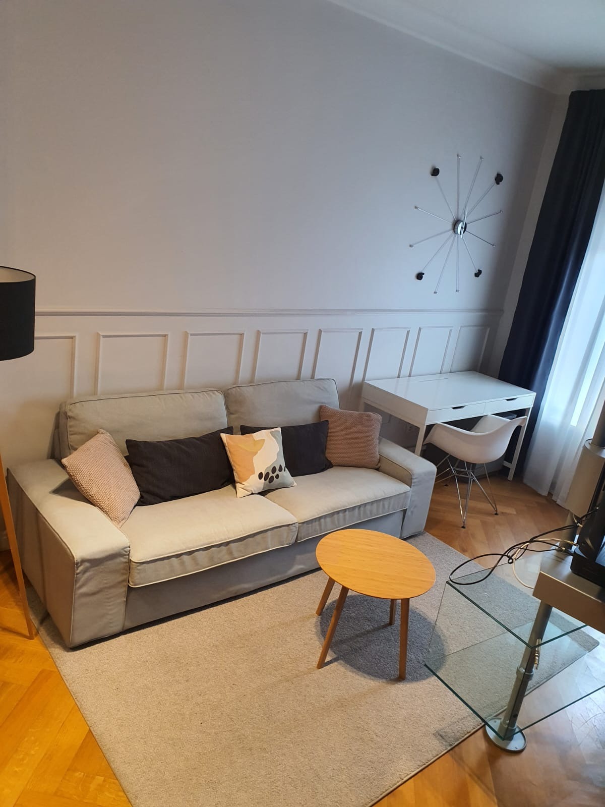 Jasnoszara sofa z poduszkami w salonie, okrągły drewniany stolik kawowy, biurko z krzesłem i dekoracyjny zegar na ścianie z boazerią.