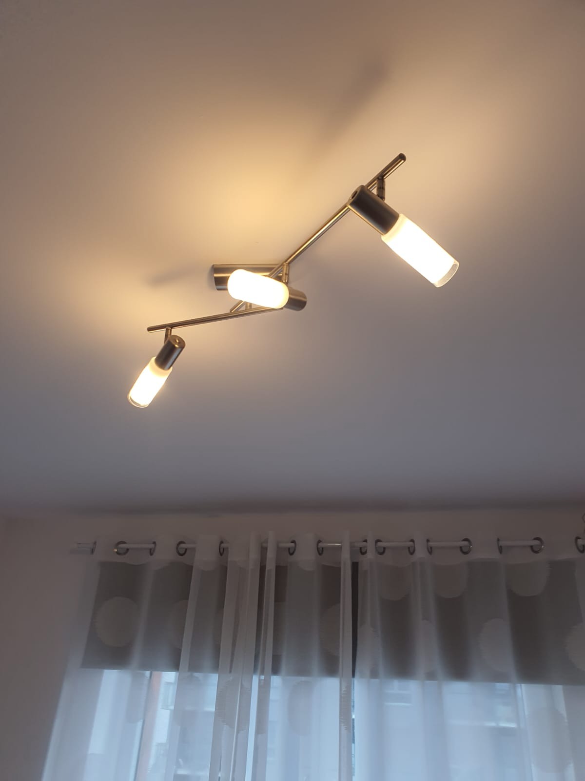 Chromowana lampa sufitowa z trzema regulowanymi reflektorami o cylindrycznym kształcie, oświetlająca pomieszczenie z delikatną firanką w tle.